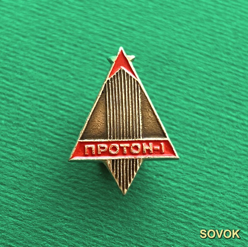 May include: A triangular metal pin featuring a red enamel top and a horizontal red enamel bar across the center. The bar displays the Cyrillic text PROTON-1 in raised gold lettering. The remaining surface has a textured gold vertical line pattern.