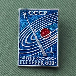 May include: A rectangular blue and silver pin with the text "CCCP" at the top. The pin features a star, a red circle, and curved lines, with the text "ИНТЕРКОСМОС-КОПЕРНИК 500" at the bottom. The pin is on a green surface.