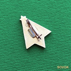 May include: A star-shaped metal pin with a textured silver-tone finish. The back features a simple wire pin mechanism with a small metal clasp attached to the center. The surface of the pin has a slightly irregular, matte metallic appearance.