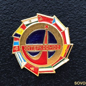 May include: A gold-colored Interkosmos pin with a central blue and red design featuring a stylized rocket. The pin is surrounded by a ring of colorful flags and the Cyrillic text "Интеркосмос".