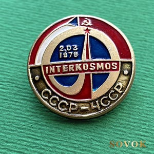 May include: A circular metal pin featuring a red, white, and blue enamel design. The center displays a rocket graphic, the text INTERKOSMOS, the date 2.03 1978, a hammer and sickle symbol, and the Cyrillic text CCCP-ЧССР along the bottom edge.