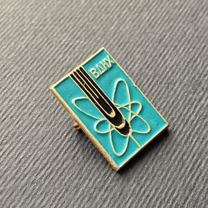 Peut inclure: &Eacute;pinglette rectangulaire avec un fond turquoise et une bordure dor&eacute;e. Elle pr&eacute;sente un motif d'atome noir et or et les lettres cyrilliques "ВДНХ" en blanc. Objet de collection vintage.