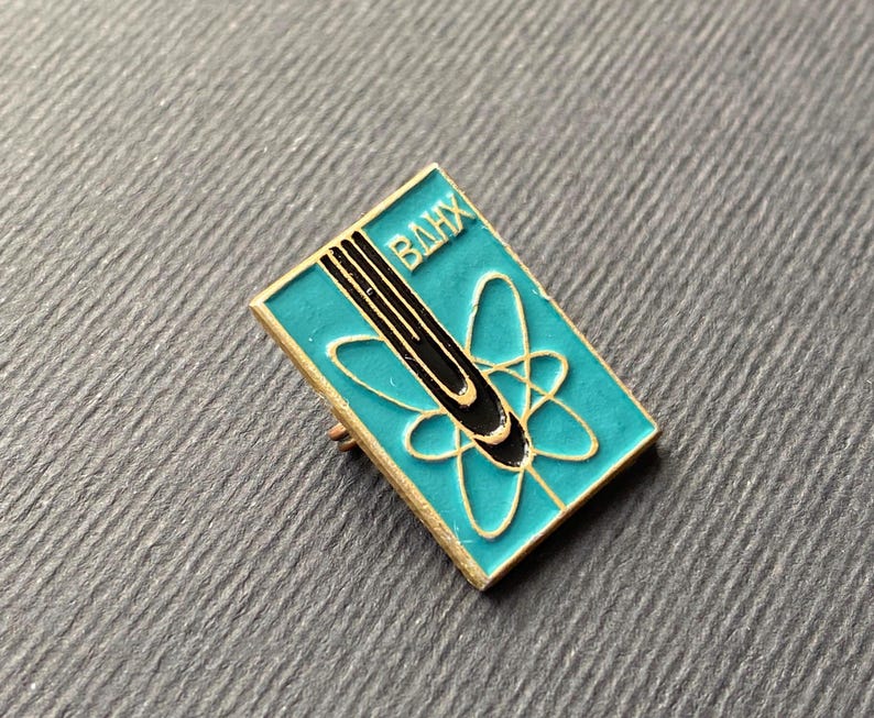 Peut inclure: &Eacute;pinglette rectangulaire avec un fond turquoise et une bordure dor&eacute;e. Elle pr&eacute;sente un motif d'atome noir et or et les lettres cyrilliques "ВДНХ" en blanc. Objet de collection vintage.