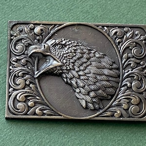 Könnte beinhalten: Eine silberfarbene Gürtelschnalle mit einem Adlermotiv. Der Adler ist im Profil dargestellt, mit dem Kopf nach links gedreht. Die Schnalle ist von einem dekorativen Rand aus Schnörkeln und Blättern umgeben.