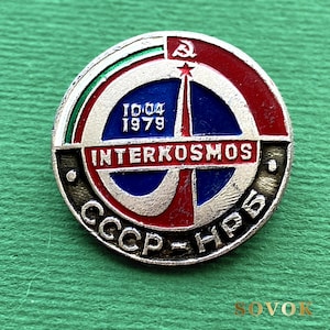 May include: A circular metal pin featuring a red, white, and blue design with a central star and rocket motif. The text reads INTERKOSMOS, 10.04 1979, and CCCP-НРБ. A hammer and sickle symbol is at the top, with metallic silver accents throughout.