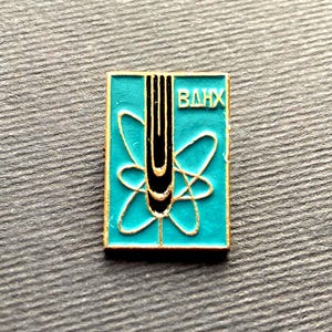 Peut inclure: Une &eacute;pingle carr&eacute;e en &eacute;mail turquoise avec une bordure dor&eacute;e. L'&eacute;pingle pr&eacute;sente un design d'atome stylis&eacute; avec une bande verticale noire et or et les lettres cyrilliques "ВДНХ" en blanc.