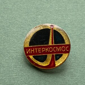 May include: A round, vintage Interkosmos pin with a gold-colored border. The pin features a black center with a red and gold design and the word "Interkosmos" in red Cyrillic script. The pin is likely a collectible item.