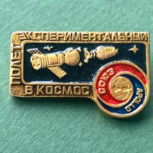 May include: Vintage space exploration pin with gold-colored accents. The pin features a spacecraft design and Cyrillic text, with a red, white, and blue circular emblem that reads "Apollo" and "Soyuz".