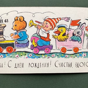 May include: A vintage birthday card featuring a colorful toy train with a teddy bear, a boy in a red and white hat, a duck, a rabbit, and a pear-shaped character. The train is on a green track with white flowers. The card says "Galushka! With a birthday! Happiness, health, success!" and the date "24.XI.65."
