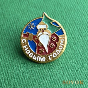 以下が含まれることがあります： 金色の金属製の丸いピンバッジ。中央には赤いコートを着て白い髭と帽子をかぶったジェド・マロースが描かれています。背景には青と緑のエナメル部分があり、金色の雪の結晶のディテールとキリル文字で「C HOBЫM ГОДОМ!」と記されています。