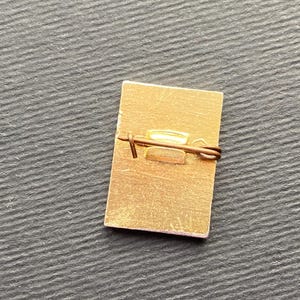 Peut inclure: Une &eacute;pingle rectangulaire de couleur or avec un fermoir de s&eacute;curit&eacute;. L'&eacute;pingle a une surface l&eacute;g&egrave;rement textur&eacute;e et un design simple. L'&eacute;pingle est pos&eacute;e sur un fond sombre et textur&eacute;.