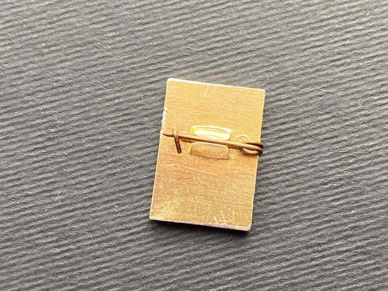 Peut inclure: Une &eacute;pingle rectangulaire de couleur or avec un fermoir de s&eacute;curit&eacute;. L'&eacute;pingle a une surface l&eacute;g&egrave;rement textur&eacute;e et un design simple. L'&eacute;pingle est pos&eacute;e sur un fond sombre et textur&eacute;.