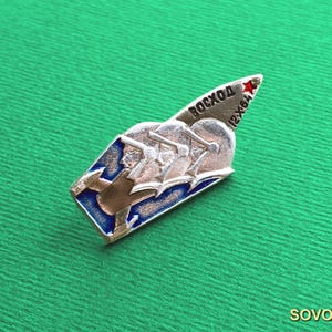 Puede incluir: Un pin de solapa de metal plateado con forma de cohete, que presenta tres cascos de astronauta estilizados en relieve, detalles en esmalte azul y el texto en cirílico "BOCXOD" junto a "12-X-64" y una pequeña estrella roja en la punta.