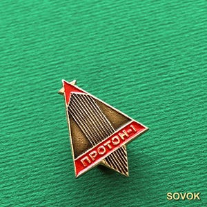 May include: A triangular metal pin featuring a red star at the top and a red border with the Cyrillic text PROTON-1 in white. The center of the pin has a dark bronze finish with vertical lines, creating a stylized rocket or spacecraft design.