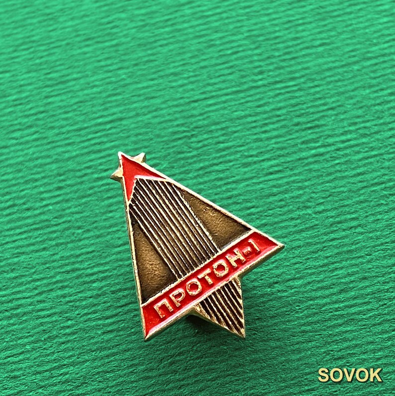 May include: A triangular metal pin featuring a red star at the top and a red border with the Cyrillic text PROTON-1 in white. The center of the pin has a dark bronze finish with vertical lines, creating a stylized rocket or spacecraft design.