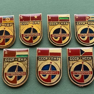 May include: A collection of seven vintage Interkosmos pins. Each pin features a gold-colored border, a red and blue design, and the text "INTERKOSMOS". The pins also display flags and Cyrillic text, likely representing different countries.