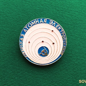 Può includere: Una spilla metallica circolare con centro bianco, anelli concentrici e piccoli punti rossi. Il bordo esterno blu contiene il testo cirillico "РОВЕНСКАЯ АТОМНАЯ ЭЛЕКТРОСТАНЦИЯ". Un piccolo cerchio blu al centro mostra un emblema dorato.