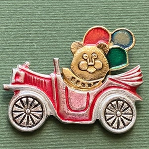 May include: A whimsical pin featuring a gold-colored bear driving a red and silver antique car. The bear is holding colorful balloons. The pin has a detailed design with a vintage aesthetic, perfect for adding a touch of charm to any outfit.