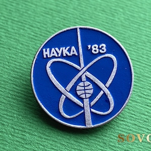 May include: A circular blue metal pin featuring a silver atomic symbol design in the center. The text "HAYKA" is printed on the left side and "'83" is printed on the right side. A small globe icon is positioned at the base of the atomic orbits.