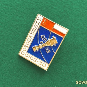 May include: A rectangular metal pin featuring a blue background with a gold-toned spacecraft illustration. The top right corner displays red and white stripes with a small gold hammer and sickle. The text reads "SOYUZ-30" in white capital letters.