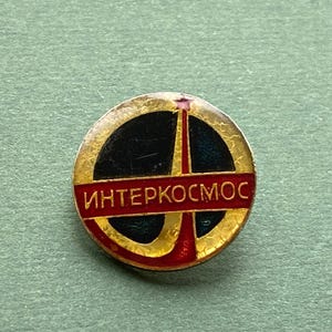 May include: A round, vintage pin with a gold-colored border and a black center. The pin features a red horizontal bar with the Cyrillic text "ИНТЕРКОСМОС" and a gold-colored tower design. The pin is set against a green background.