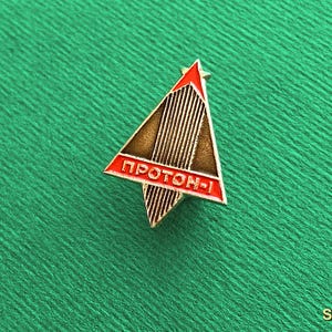 May include: A triangular metal pin featuring a stylized rocket design with vertical lines. The pin has a red border and a red horizontal bar at the base with the Cyrillic text PROTON-1 in white lettering. The central section is a metallic bronze color.