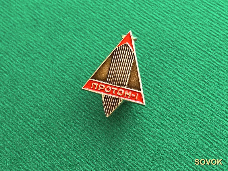 May include: A triangular metal pin featuring a stylized rocket design with vertical lines. The pin has a red border and a red horizontal bar at the base with the Cyrillic text PROTON-1 in white lettering. The central section is a metallic bronze color.