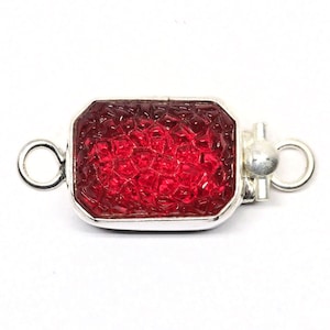 Pode incluir: Um fecho de prata com uma inserção de vidro vermelho facetado. O fecho tem dois laços de cada lado para prender a um colar ou pulseira.