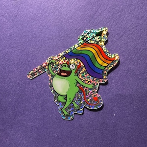 May include: A green frog with a devil tail holding a rainbow pride flag on a stick. The frog is wearing a red crown and has a mischievous expression. The background is a sparkly, iridescent material.