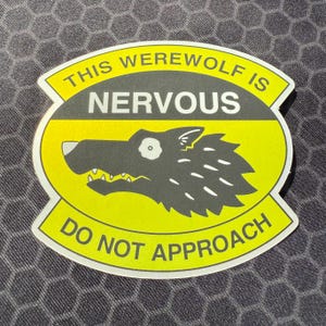 Peut inclure: Un autocollant jaune et noir avec le texte "THIS WEREWOLF IS NERVOUS DO NOT APPROACH". L'autocollant représente un loup de dessin animé avec une expression inquiète.