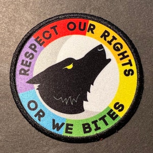 May include: A round, black and white patch with a rainbow border. The patch features a black wolf silhouette howling with a yellow eye. The text on the patch reads "Respect Our Rights Or We Bite".