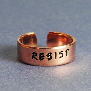 May include: A copper-colored adjustable ring with the word "RESIST" stamped in black lettering. The ring is open at the top and sits on a blue-gray surface. The ring is a simple band design.