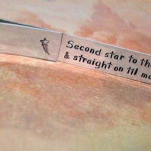 May include: A silver-toned metal tie clip with the phrase "Second star to the right & straight on til morning" engraved on it. A star and comet design is stamped on the clip. The clip is on a textured, brown surface.