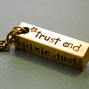 Könnte beinhalten: Eine goldfarbene Halskette mit einem rechteckigen Anhänger, auf dem "trust and pixie dust" mit einem Stern auf jeder Seite des Textes steht.
