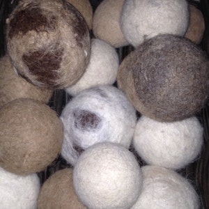 May include: A close-up of a basket filled with a variety of wool dryer balls. The balls are in shades of white, brown, and gray. Some of the balls have a speckled pattern.