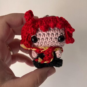 Könnte beinhalten: Eine kleine gehäkelte Puppe als Schlüsselanhänger mit leuchtend roten Haaren, einem hellrosa Gesicht und schwarzen Perlenaugen. Die Figur trägt ein schwarzes Outfit, das mit einem strukturierten rot-gelben Schal um Hals und Rumpf akzentuiert ist.