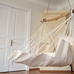 Peut inclure: Un hamac ray&eacute; blanc et beige avec un cadre en bois, suspendu au plafond dans une pi&egrave;ce aux murs blancs et une porte blanche.