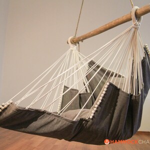 May include: A gray hammock chair with white rope and white pom-poms around the edges. The chair is hanging from a wooden bar.