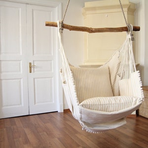 Peut inclure: Un fauteuil hamac blanc avec un cadre en bois et des coussins ray&eacute;s, suspendu dans une pi&egrave;ce avec des portes blanches et un sol en bois.