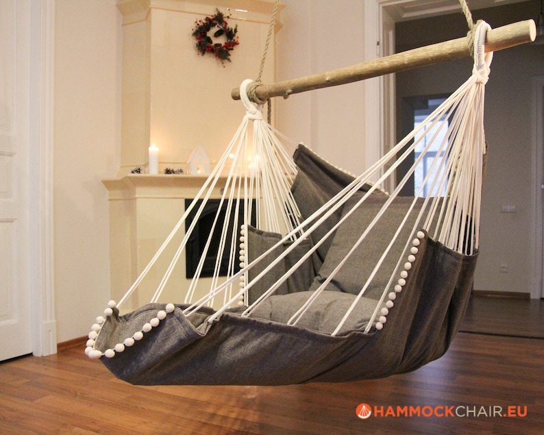 May include: A gray hammock chair with white pom-poms around the edge. The chair is hanging from a wooden beam and is in a room with a fireplace.