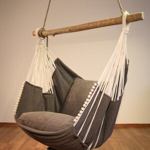 May include: A gray hammock chair with white rope and wooden spreader bar. The chair has a gray cushion and white pom-poms around the edges.