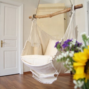 Peut inclure: Un fauteuil hamac en macram&eacute; blanc avec un coussin ray&eacute; blanc est suspendu &agrave; une poutre en bois. Le fauteuil est dans une pi&egrave;ce aux murs blancs et au sol en bois. Un bouquet de fleurs est au premier plan.