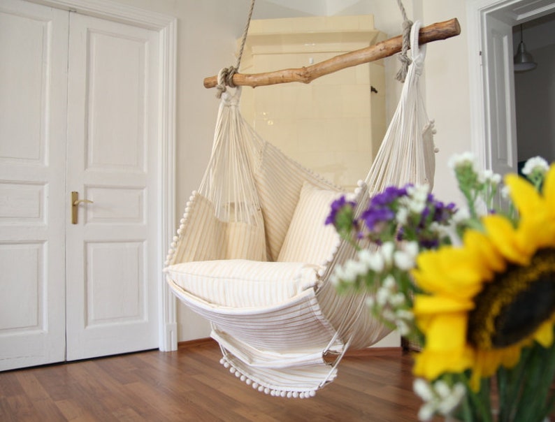 Peut inclure: Un fauteuil hamac en macram&eacute; blanc avec un coussin ray&eacute; blanc est suspendu &agrave; une poutre en bois. Le fauteuil est dans une pi&egrave;ce aux murs blancs et au sol en bois. Un bouquet de fleurs est au premier plan.