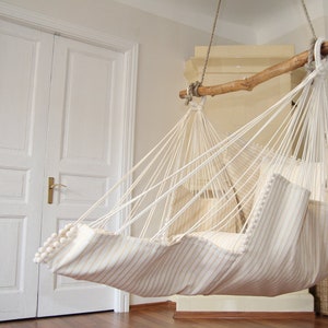Peut inclure: Un hamac ray&eacute; blanc et beige avec un cadre en bois, suspendu au plafond dans une pi&egrave;ce aux murs blancs et une porte blanche.