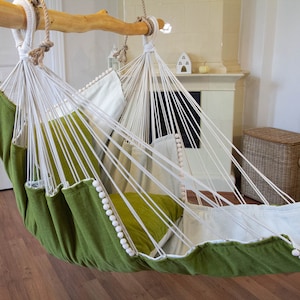 Puede incluir: Una silla hamaca verde y blanca con pompones blancos alrededor del borde. La hamaca está colgada de una barra de madera y está suspendida en una habitación con suelo de madera.