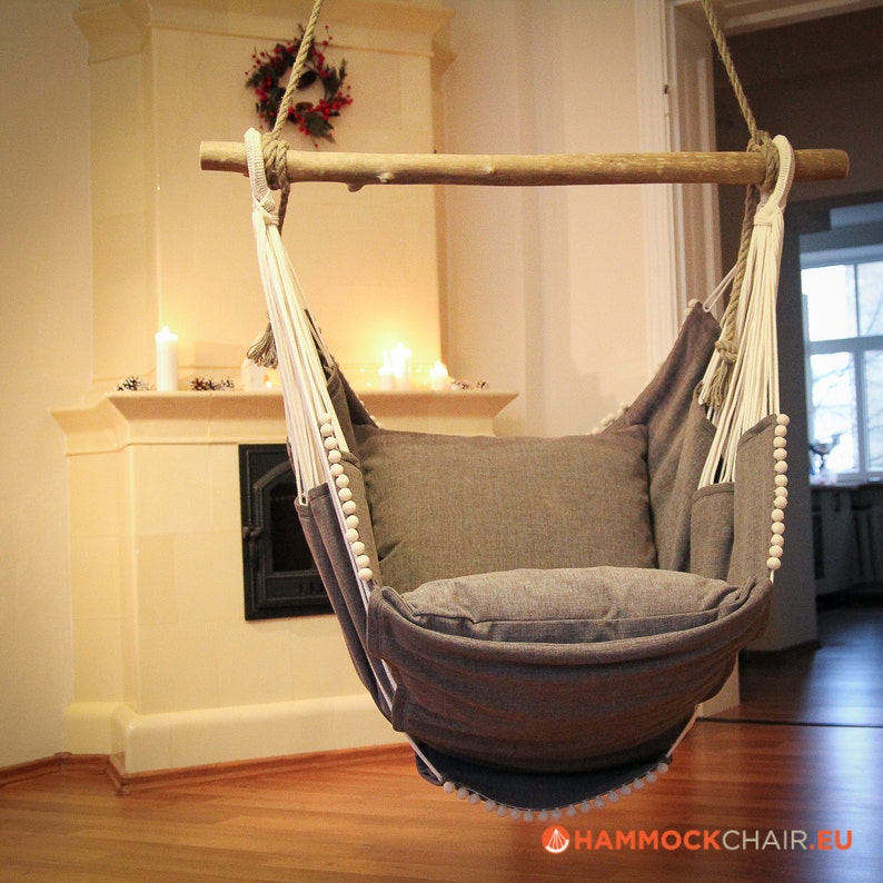 May include: A gray hammock chair with white rope and wooden support bar. The chair is hanging in a room with a fireplace in the background.