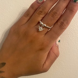 Puede incluir: Un anillo de plata con un pequeño dije de abeja colgando. El anillo está hecho de pequeñas cuentas y se usa en un dedo con uñas pintadas.
