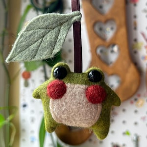 May include: A handmade felt frog ornament with a green body, red cheeks, and black eyes. The frog has a cream-colored belly and is attached to a brown ribbon with a green felt leaf. The ornament is a whimsical decoration.