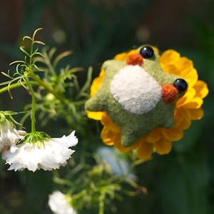 May include: A handcrafted felt frog with black eyes, orange details, and a white belly rests on a vibrant yellow flower. The frog is green and has a playful design. The background is a blurred mix of green foliage.