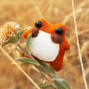 以下が含まれることがあります： 黒い目と白いお腹を持つ、手作りのオレンジ色のフェルトのカエルが、緑色の植物の茎に止まっています。カエルは茶色のアクセントがあり、乾燥した草と葉のぼやけた背景に配置されています。気まぐれな手作りのアイテムです。